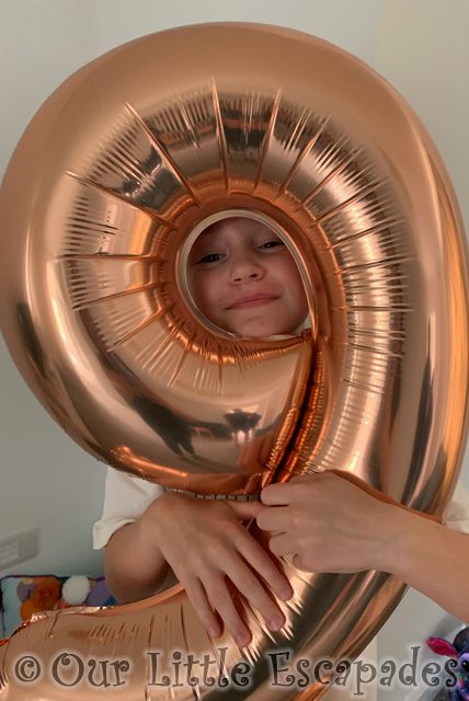 little e smiling face through rose gold number nine balloon Ninth Birthday
